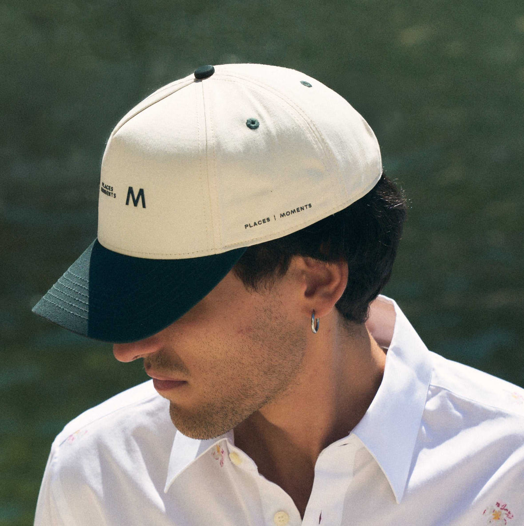The Places Moments logo baseball cap in cream with a green brim, the top button and eyelets picked out in the same green.