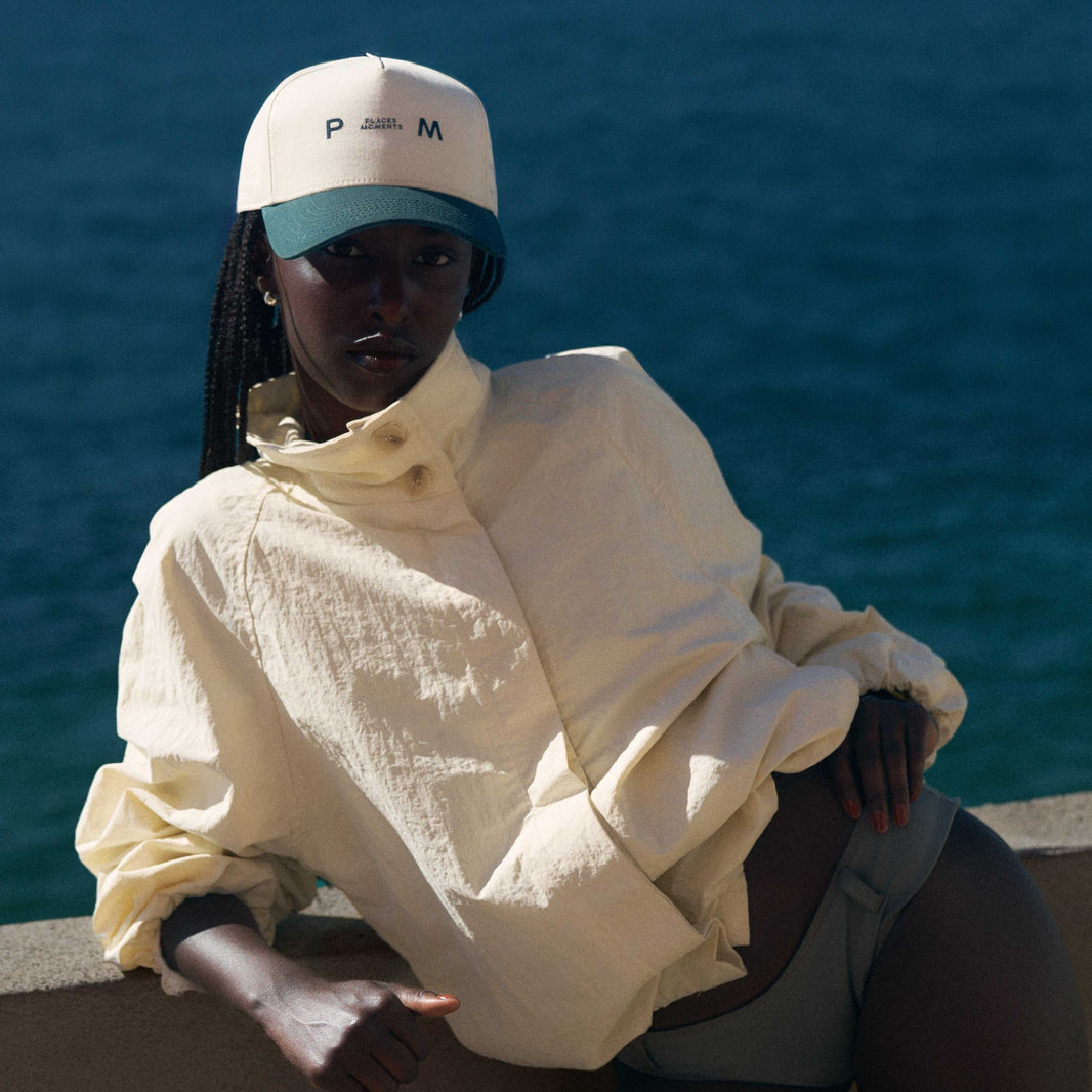 A person wearing the cream and green Places Moments logo baseball cap by the water.