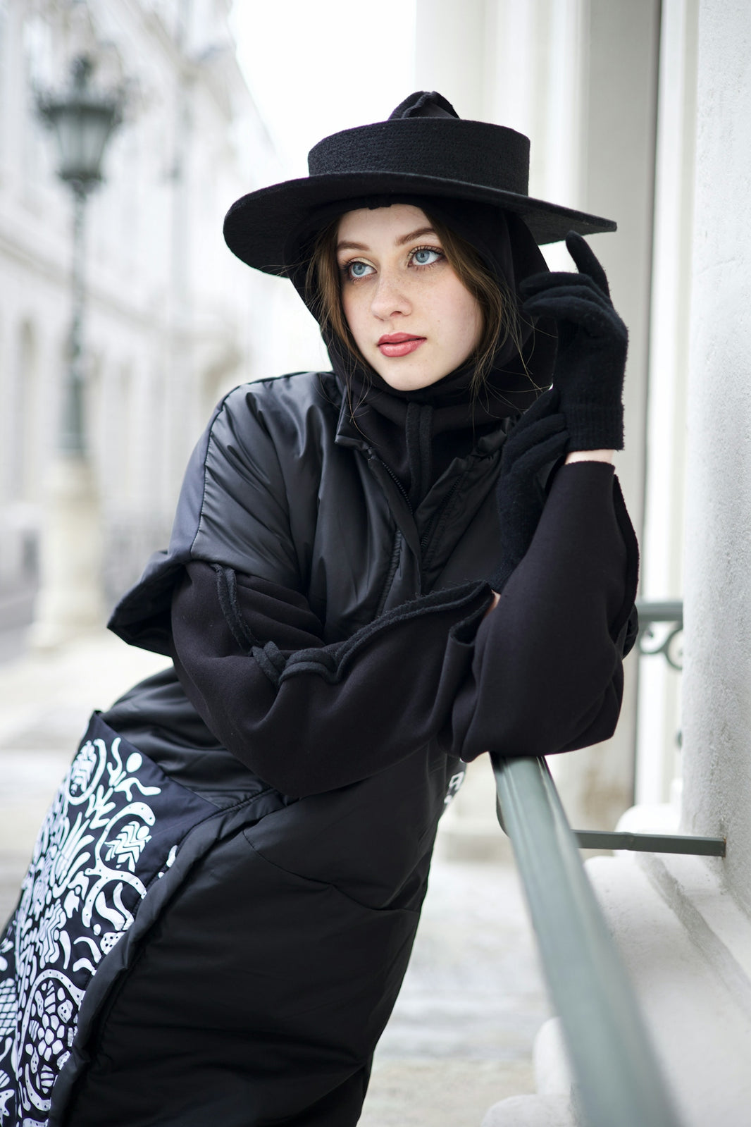 a woman in a black coat and hat leaning against a wall