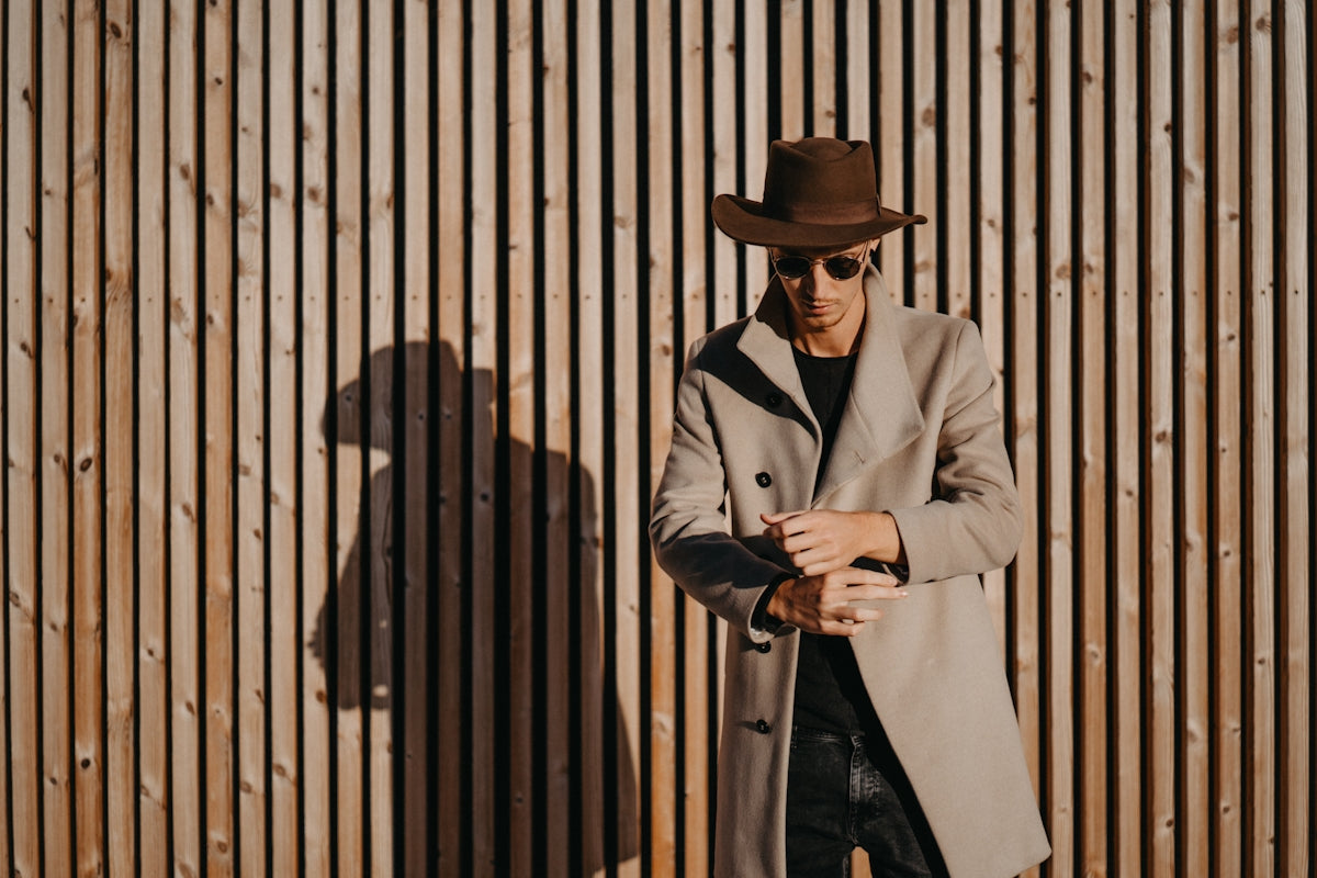 A man in a trench coat and hat leaning against a wall
