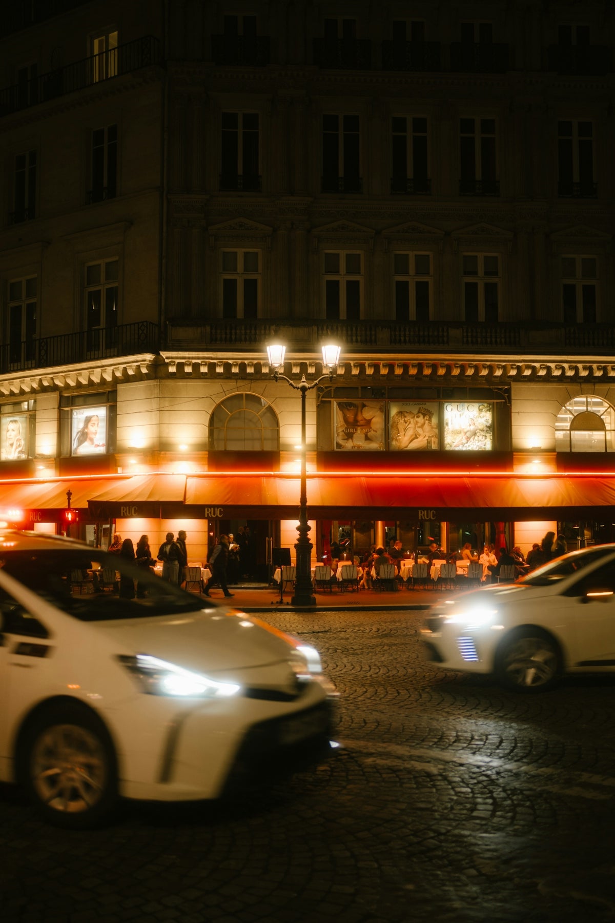 Cars passing by a brightly lit parisian cafe at night.