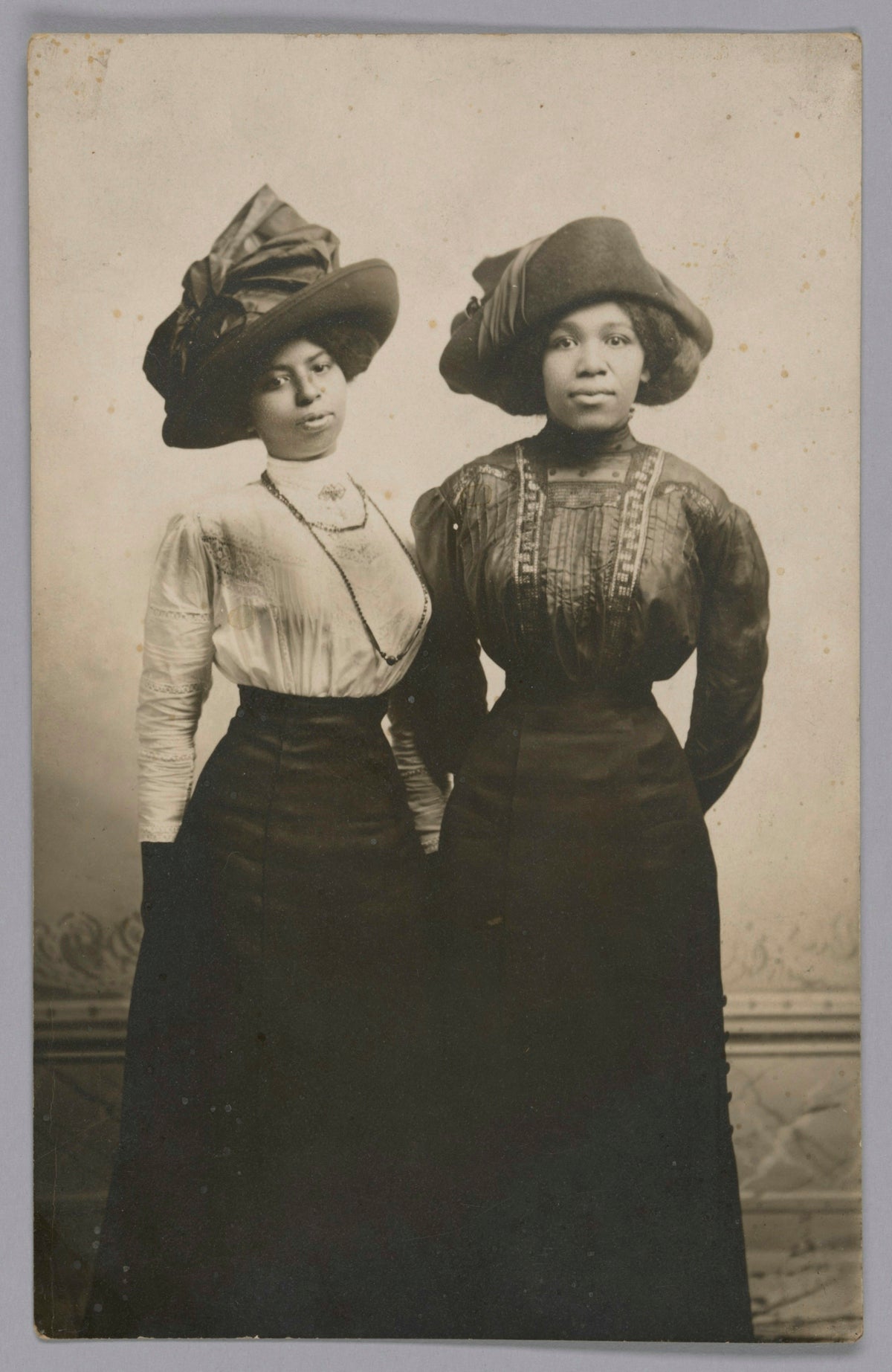 Two women wearing large hats and long dresses stand together.