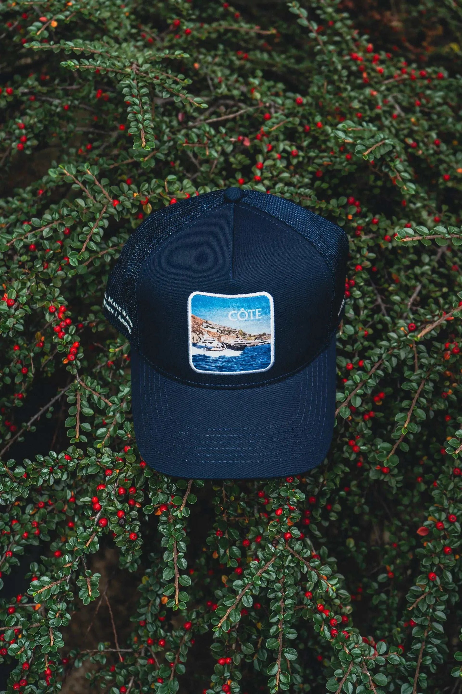Côte Mesh-Back Trucker Hat in Indie Blue with mesh back and Places | Moments patch placed on green foliage with red berries