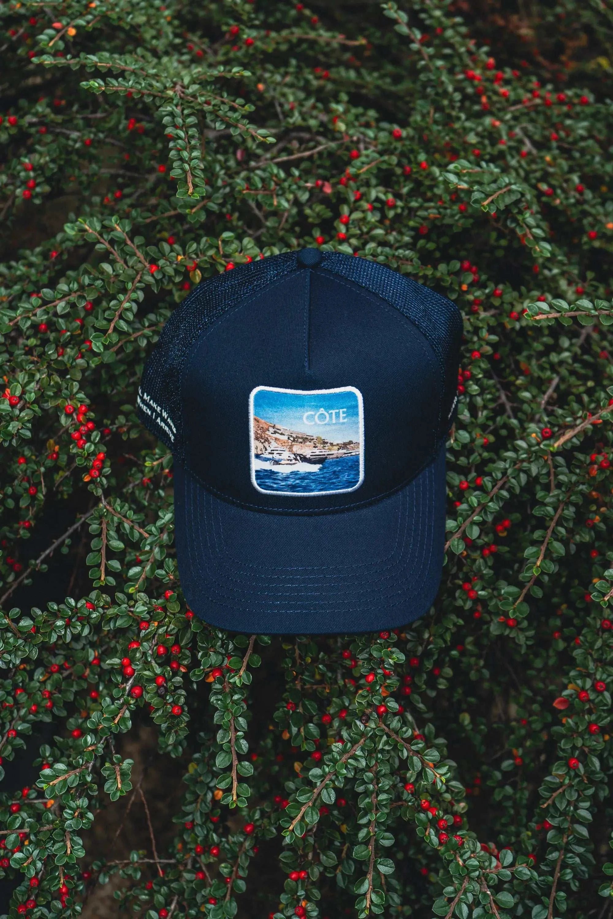 Côte Mesh-Back Trucker Hat in Indie Blue with mesh back and Places | Moments patch placed on green foliage with red berries