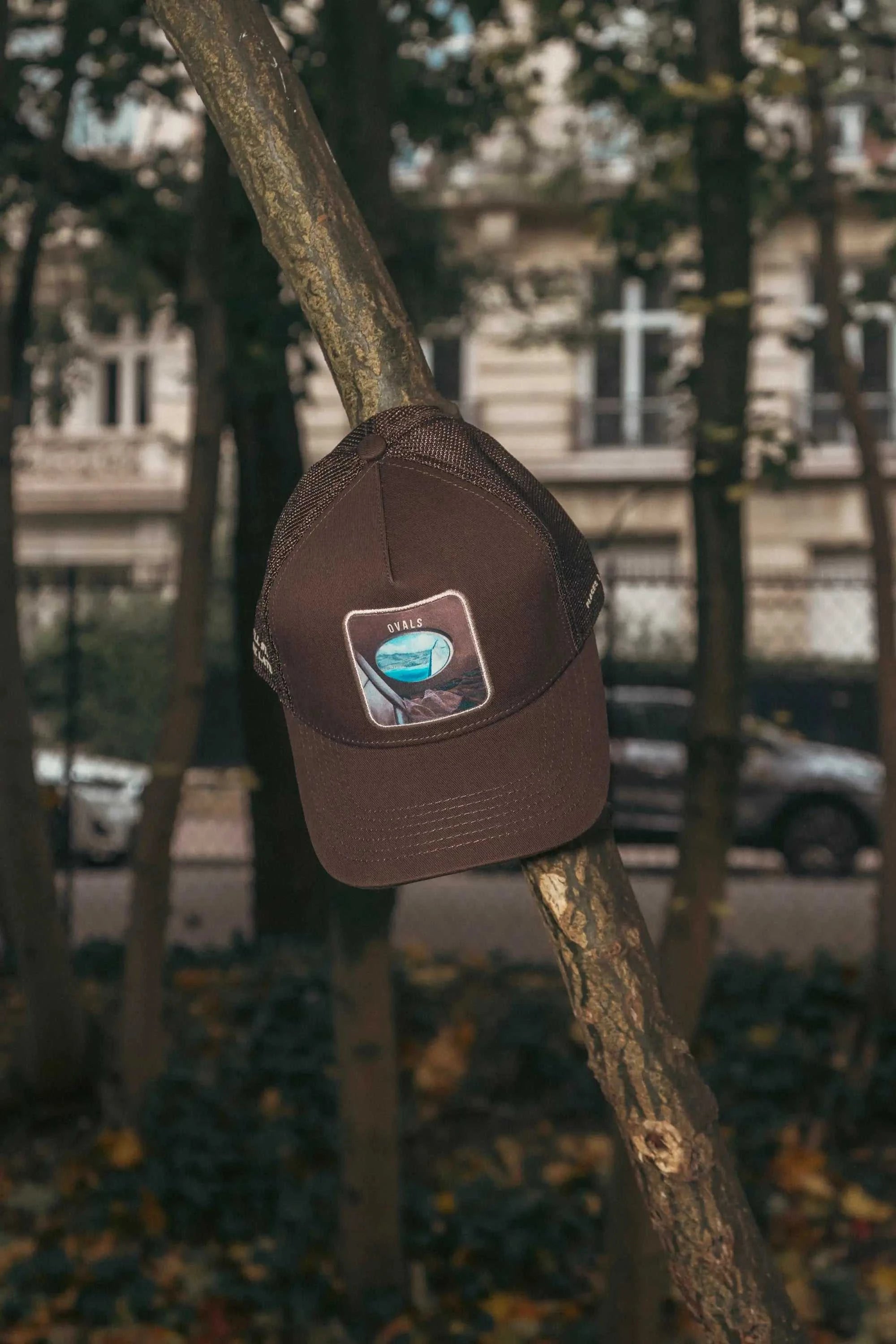 Ovals Mesh-Back Trucker Hat in chestnut brown with breathable tan mesh and embroidered patch hanging on tree branch outdoors