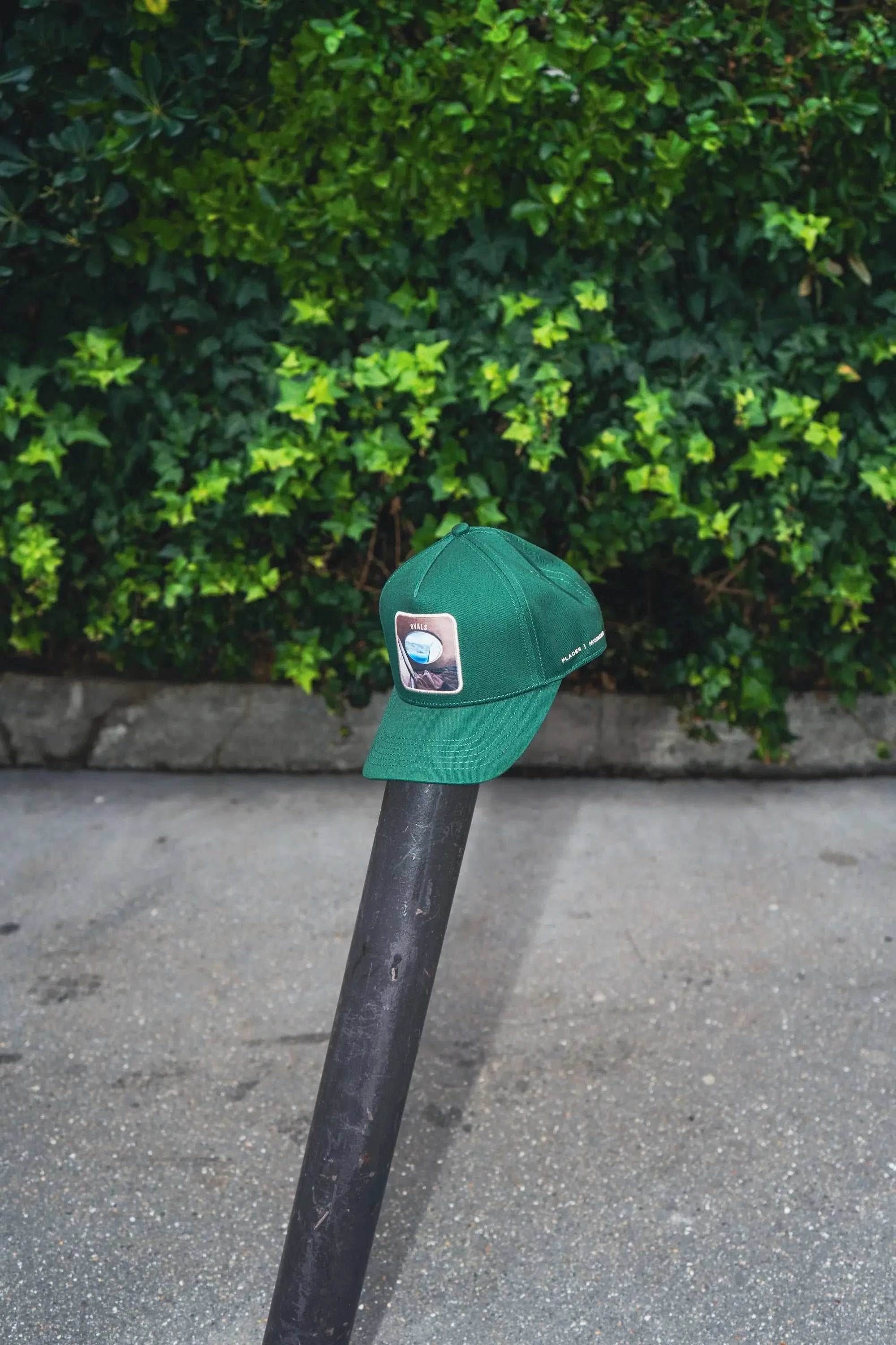 Forest green Ovals Baseball Cap with embroidered patch on a pole outdoors against leafy background