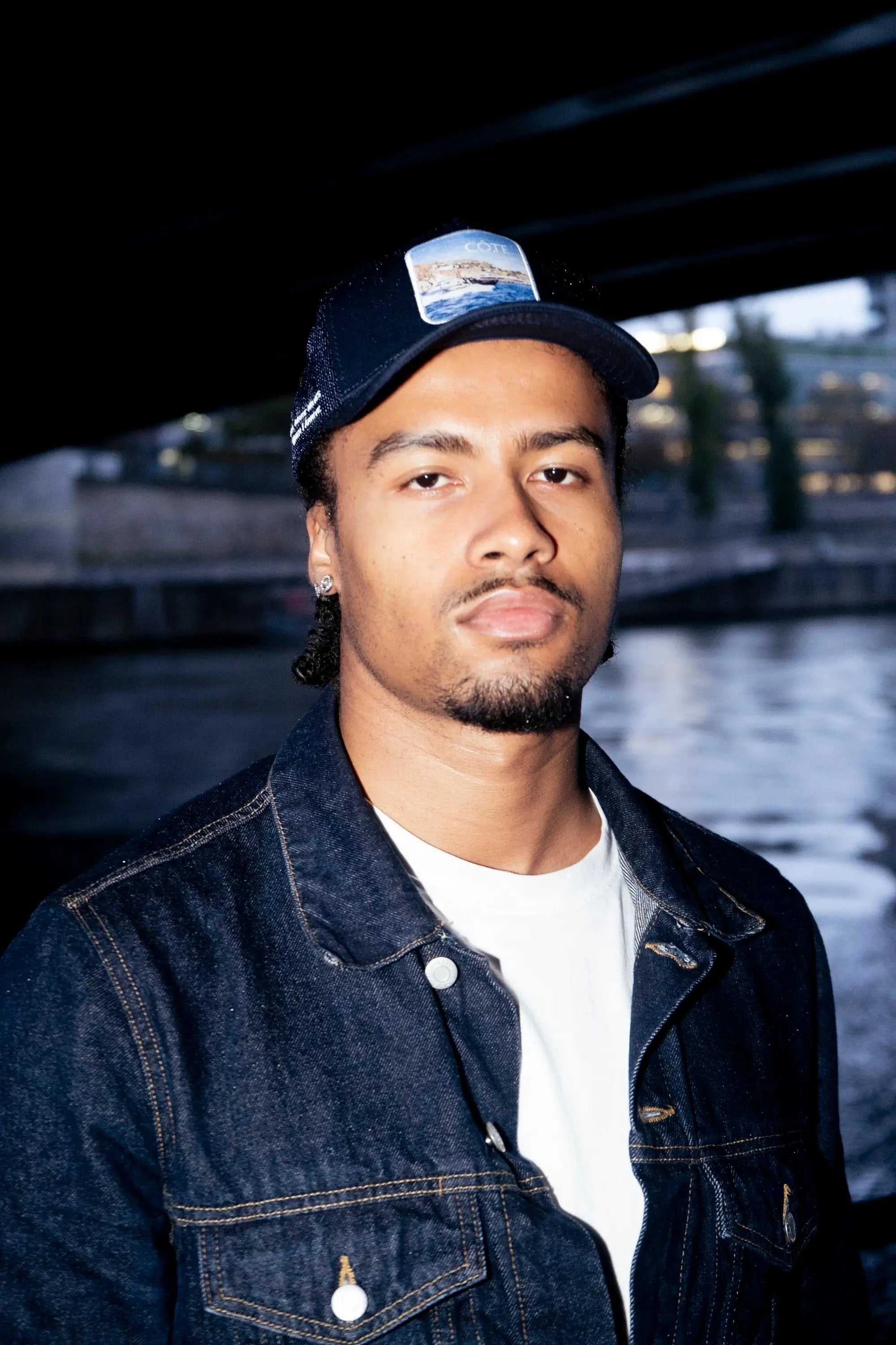 Man wearing Côte Mesh-Back Trucker Hat in Indie Blue with mesh backing and signature patch under a bridge near water