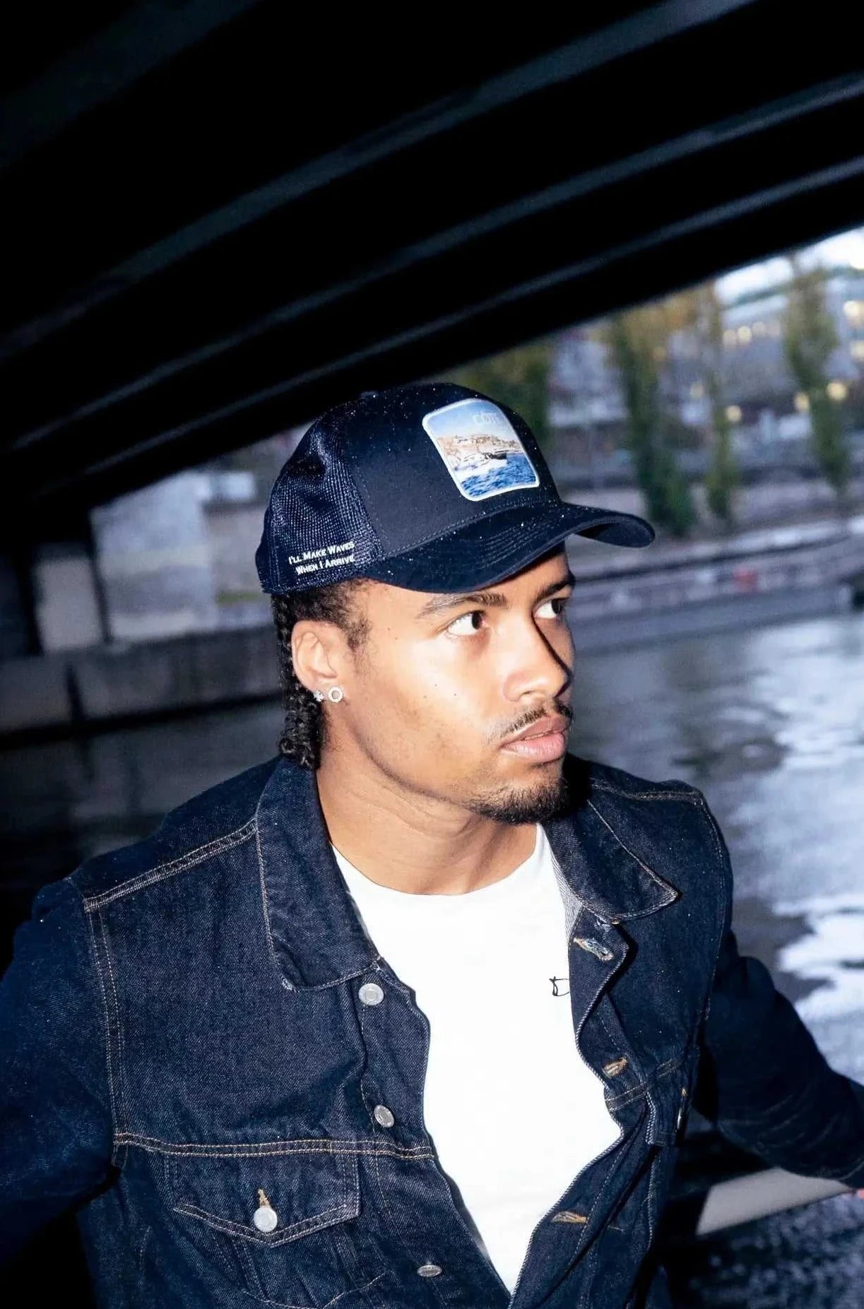 Man wearing Côte Mesh-Back Trucker Hat in Indie Blue with denim jacket near water under bridge at night
