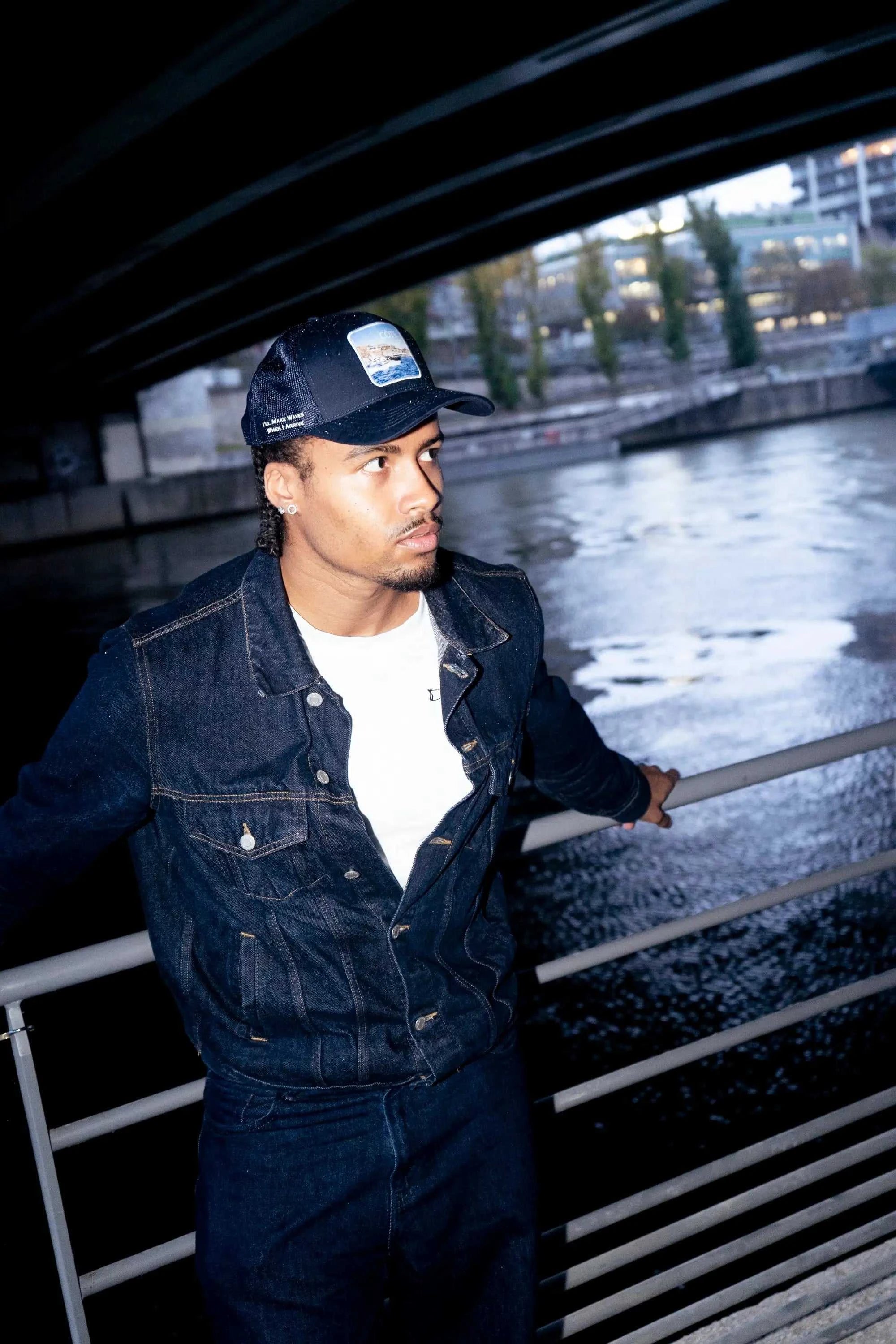 Man wearing Côte Mesh-Back Trucker Hat in Indie Blue with denim jacket near water under bridge at night