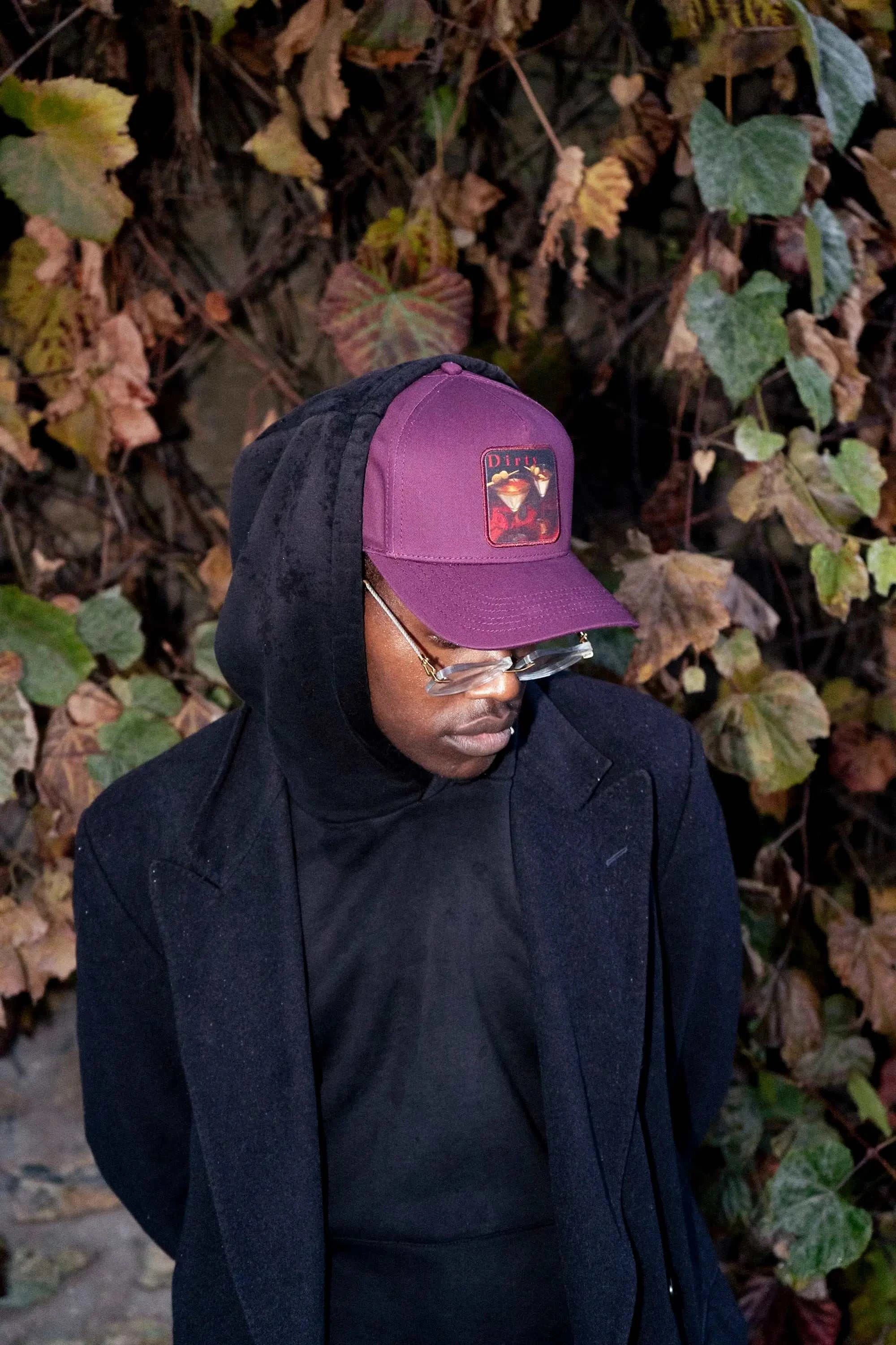 Man wearing Indian Creek Red Dirty Martini Baseball Cap with black hoodie and coat outdoors against foliage background