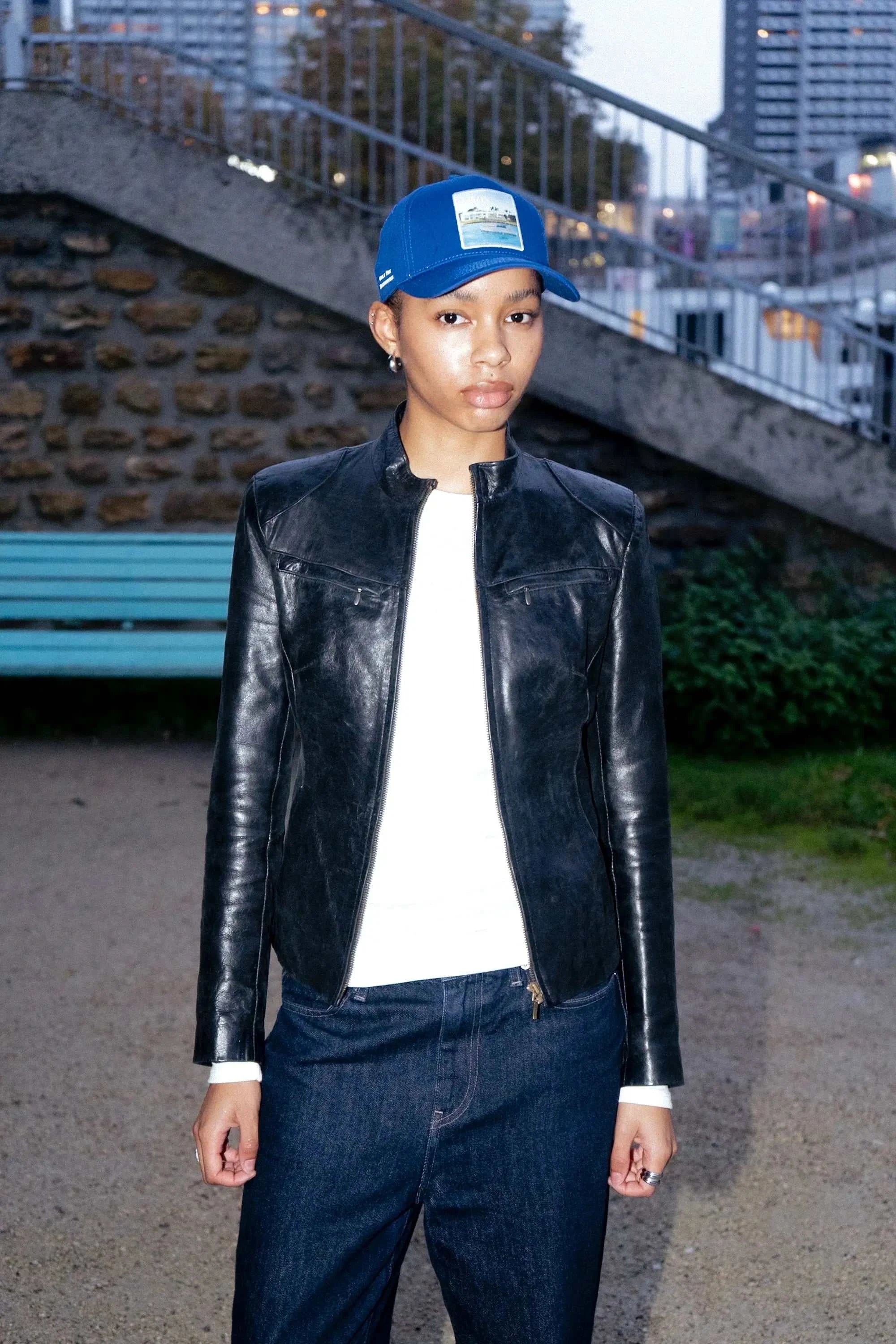Person wearing blue Nixon Baseball Cap with black leather jacket and dark jeans outdoors in urban setting