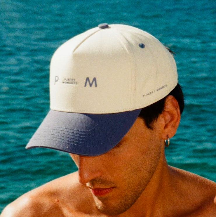 A man wearing the two-tone Places Moments logo cap, cream crown with a light blue brim and the P M logo in navy.