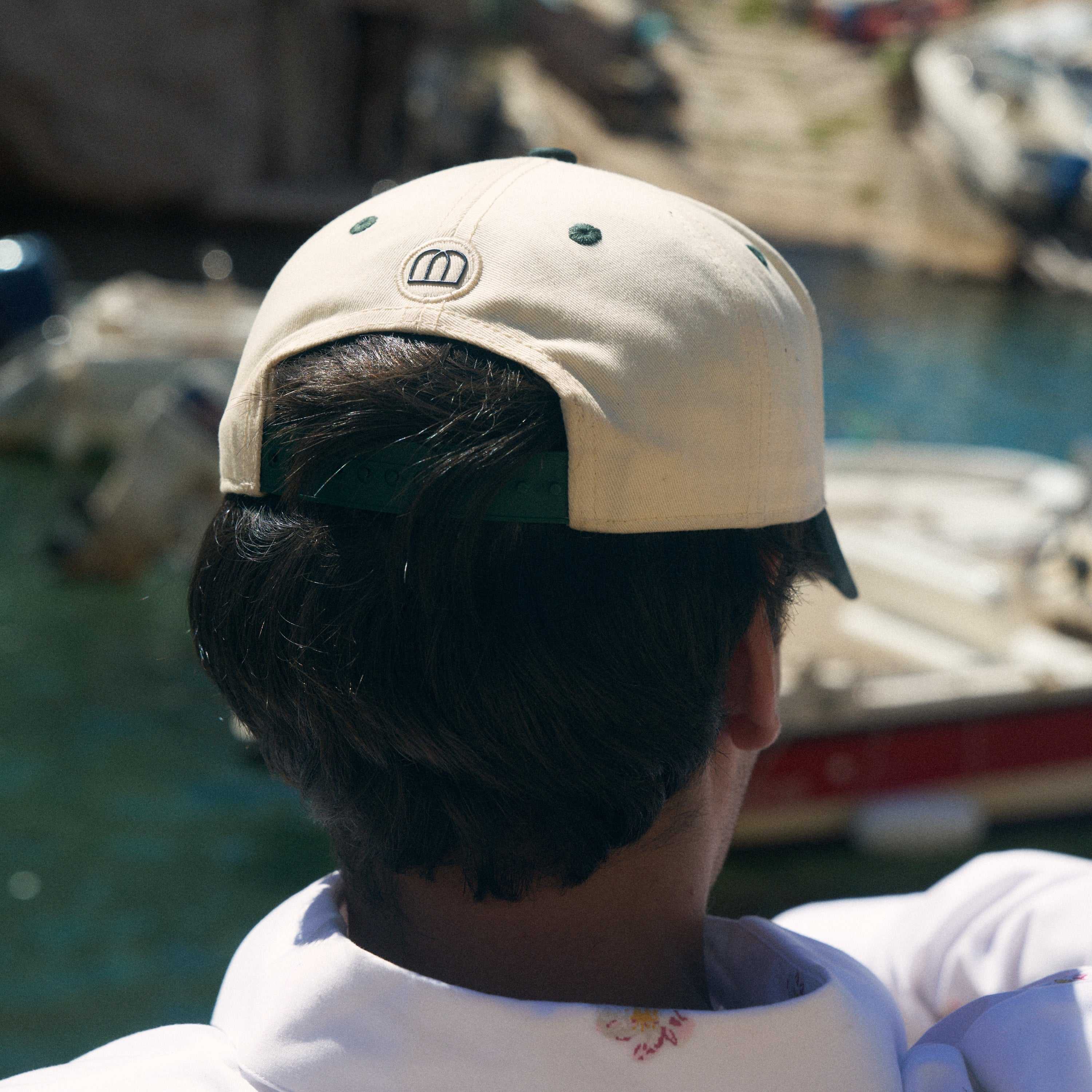 A person viewed from behind wearing a beige baseball cap with a small embroidered logo on the back, standing outdoors near water and boats.