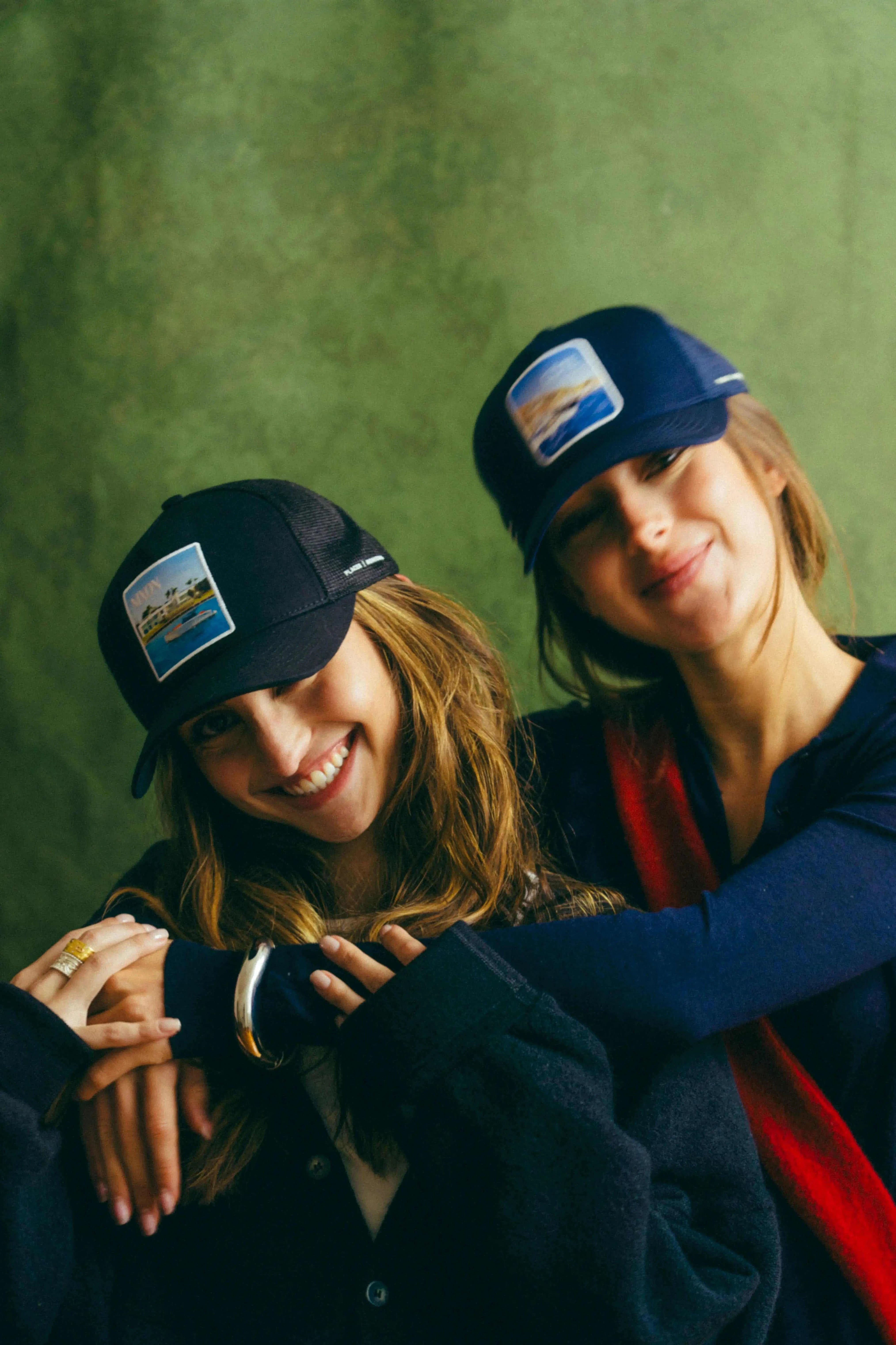 Two women wearing Nixon Mesh-Back Trucker Hats with embroidered patches and curved brims in casual outfits against a green backdrop