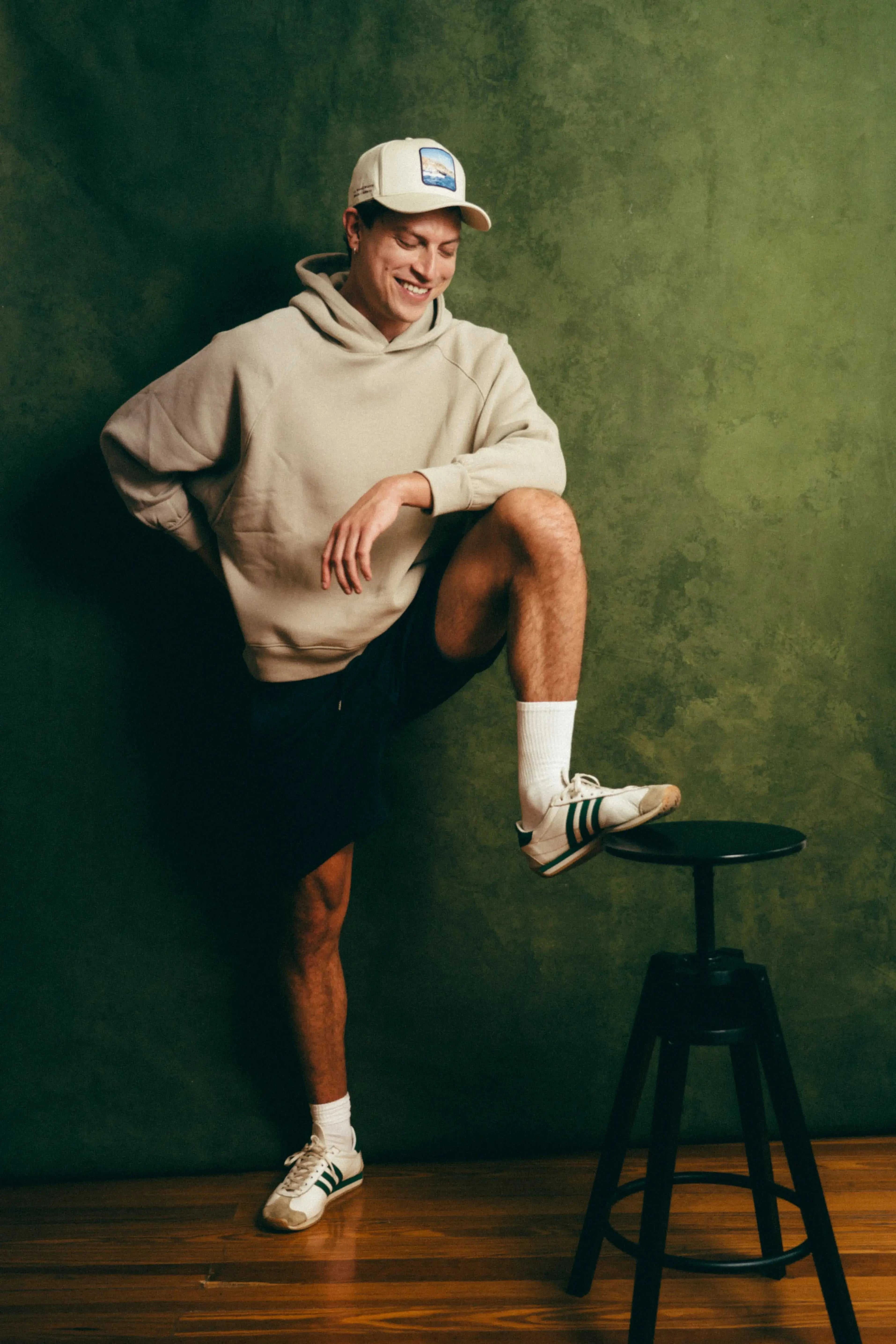 Man wearing Côte Baseball Cap in Isla Crema with beige hoodie and shorts against green background