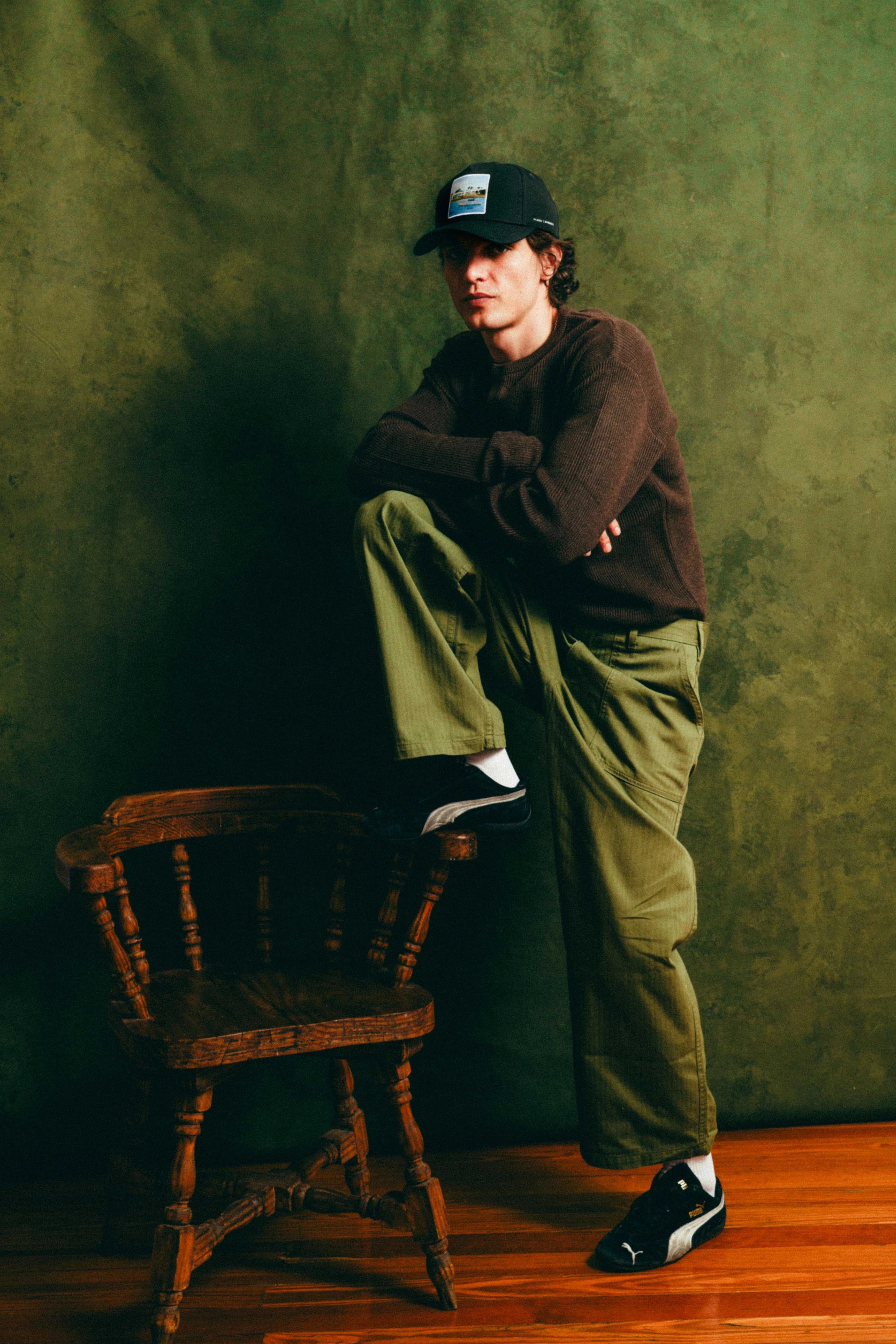 Man wearing black Nixon Mesh-Back Trucker Hat with green cargo pants and brown sweater, posing by wooden chair against green textured wall