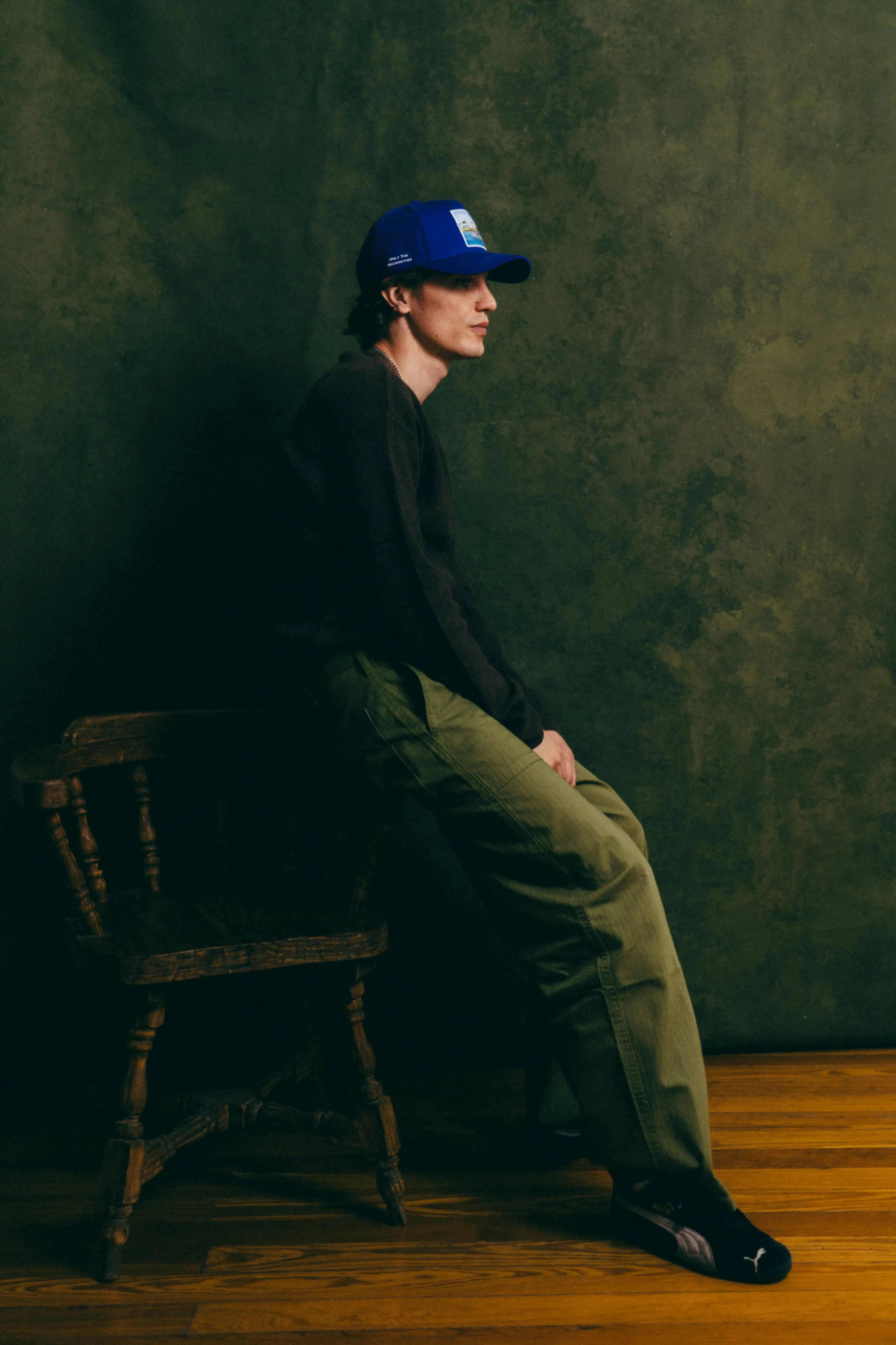 Blue Nixon Baseball Cap with structured crown and curved brim worn by man sitting on wooden chair against dark green background