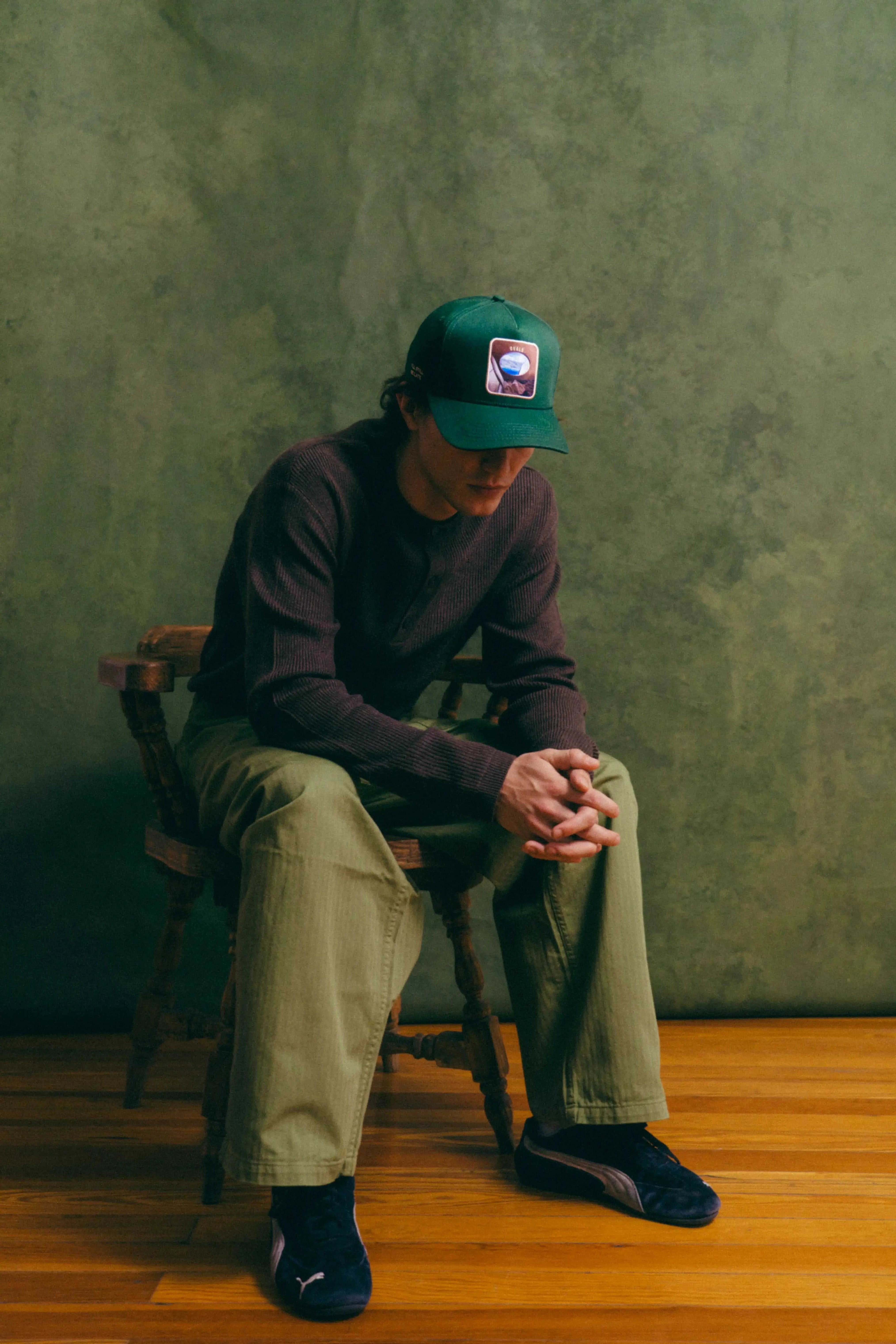 Man wearing dark green Ovals Baseball Cap with embroidered patch sitting on wooden chair