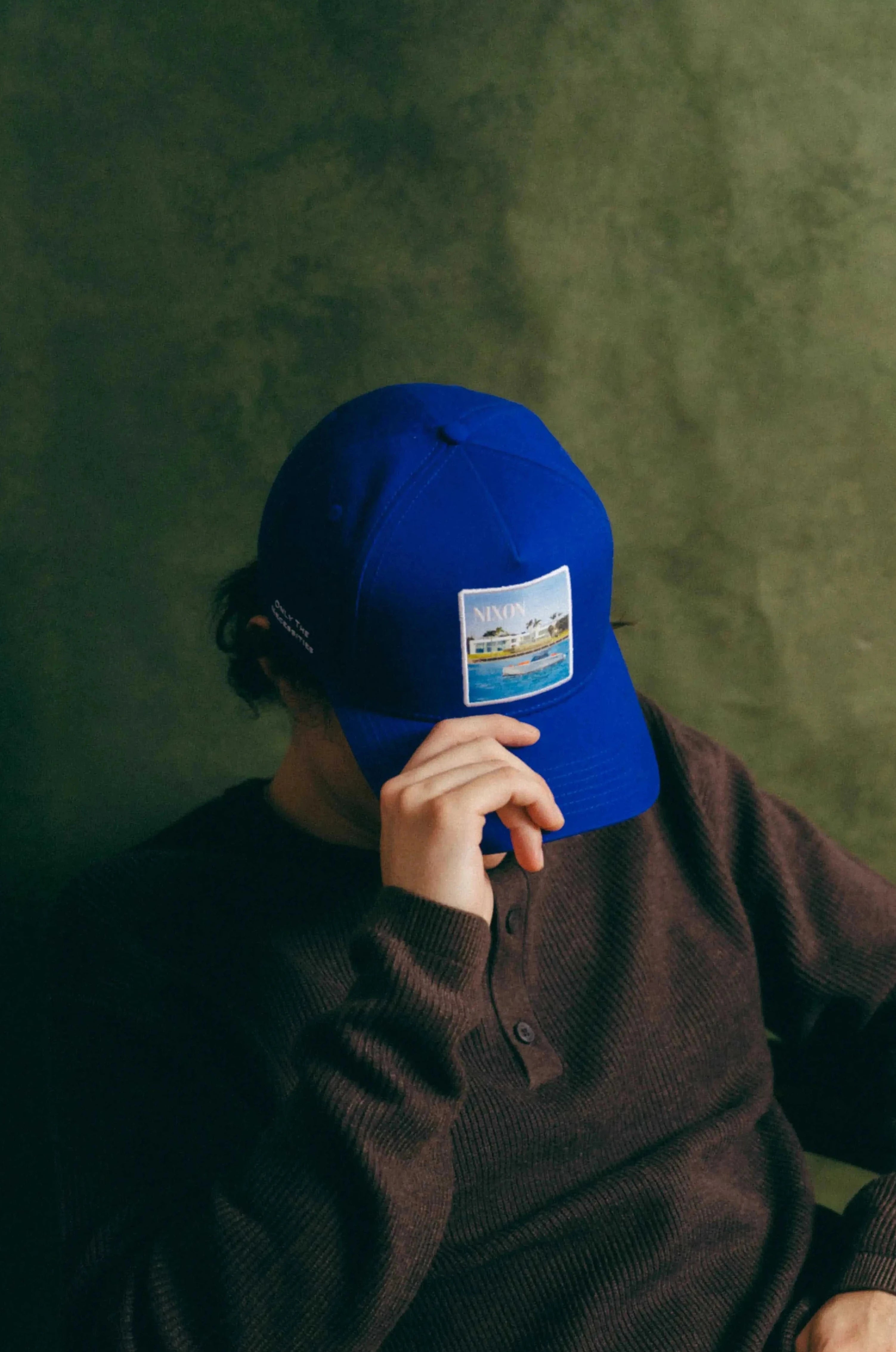 Blue Nixon Baseball Cap with curved brim and Places | Moments patch worn by person sitting in chair