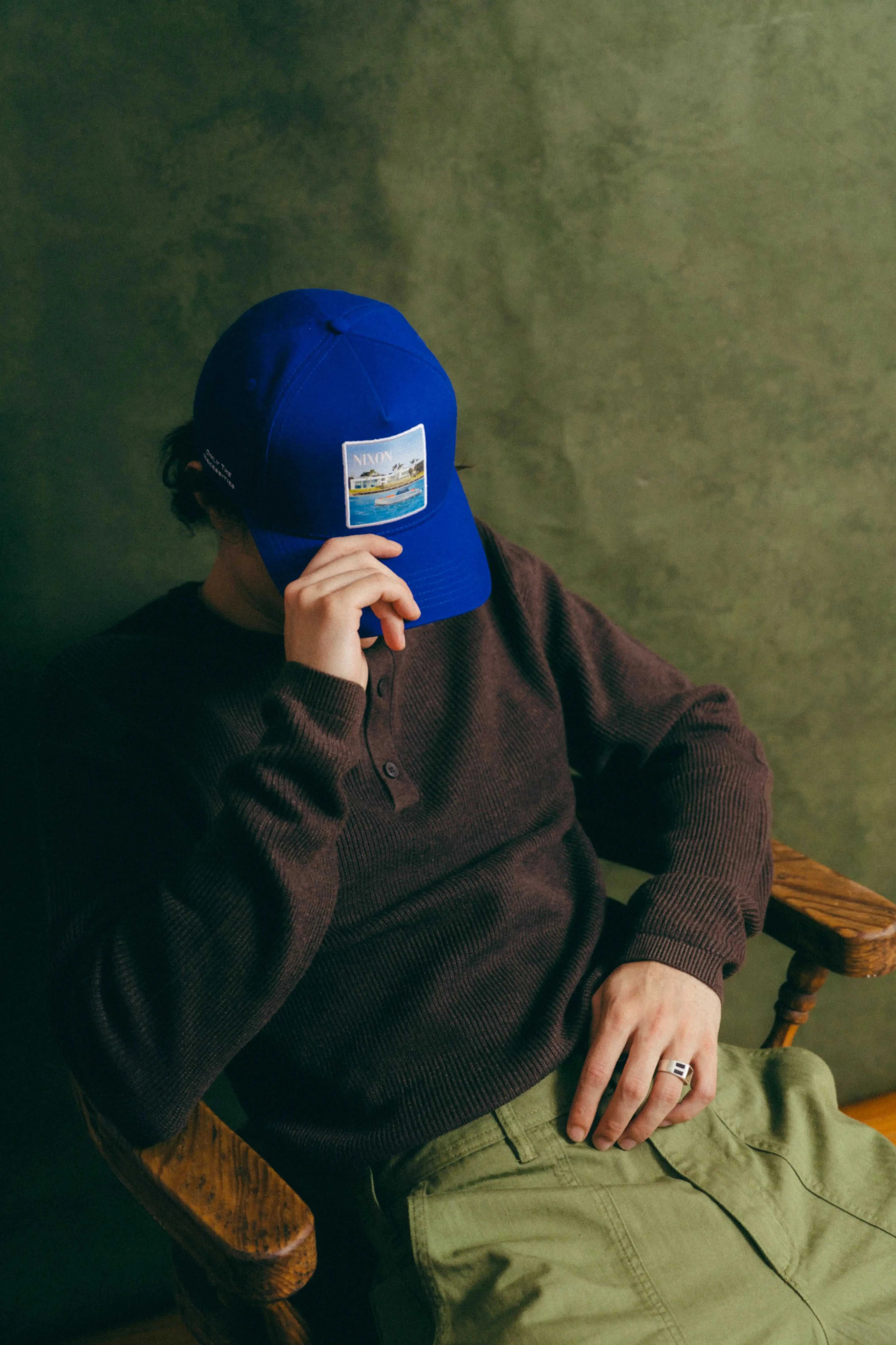 Blue Nixon Baseball Cap with curved brim and Places | Moments patch worn by person sitting in chair