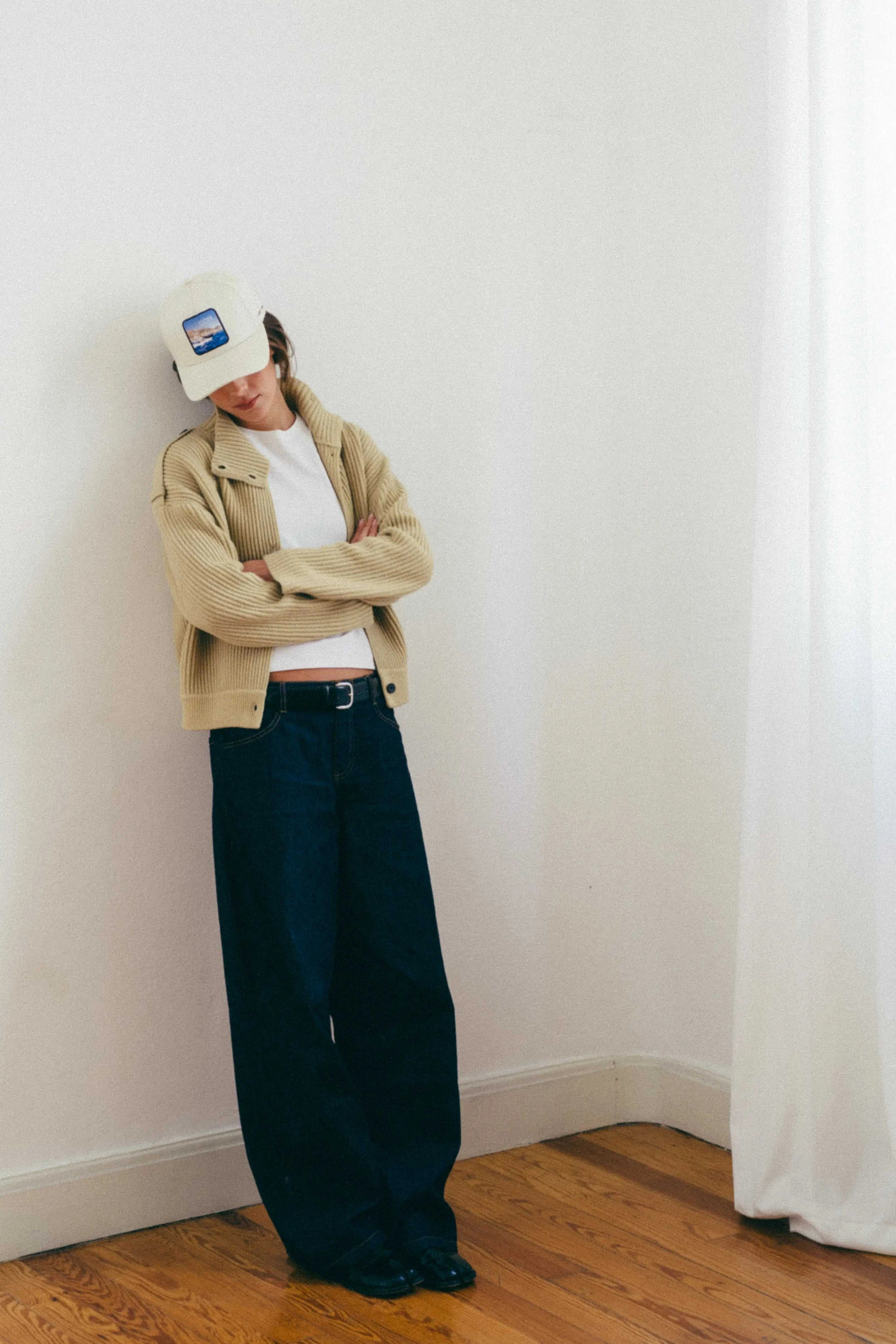Woman wearing Côte Baseball Cap in Isla Crema with casual beige jacket and dark wide-leg pants indoors