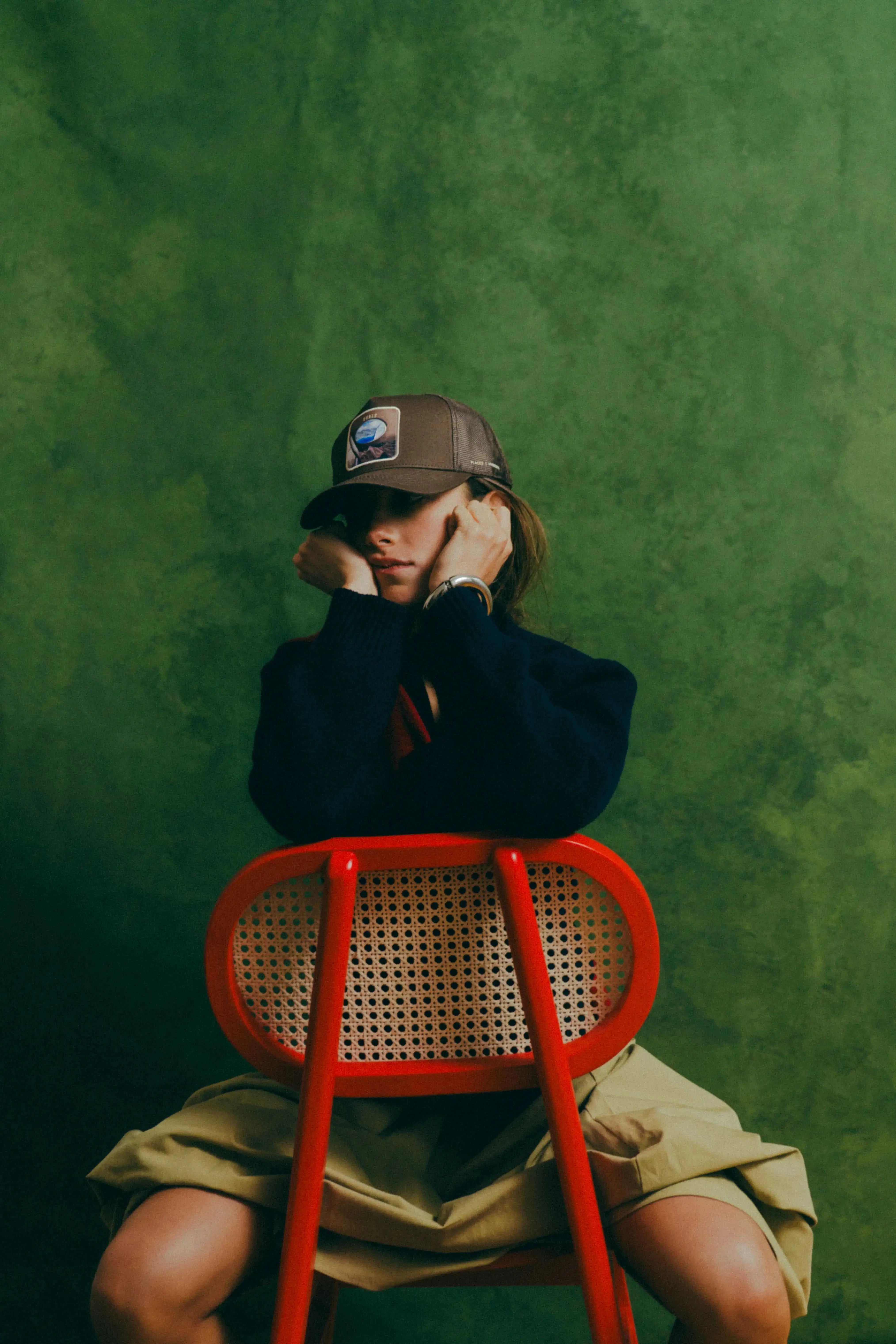 Ovals Mesh-Back Trucker Hat with chestnut front panel and tan mesh back worn by person sitting on red chair against green background