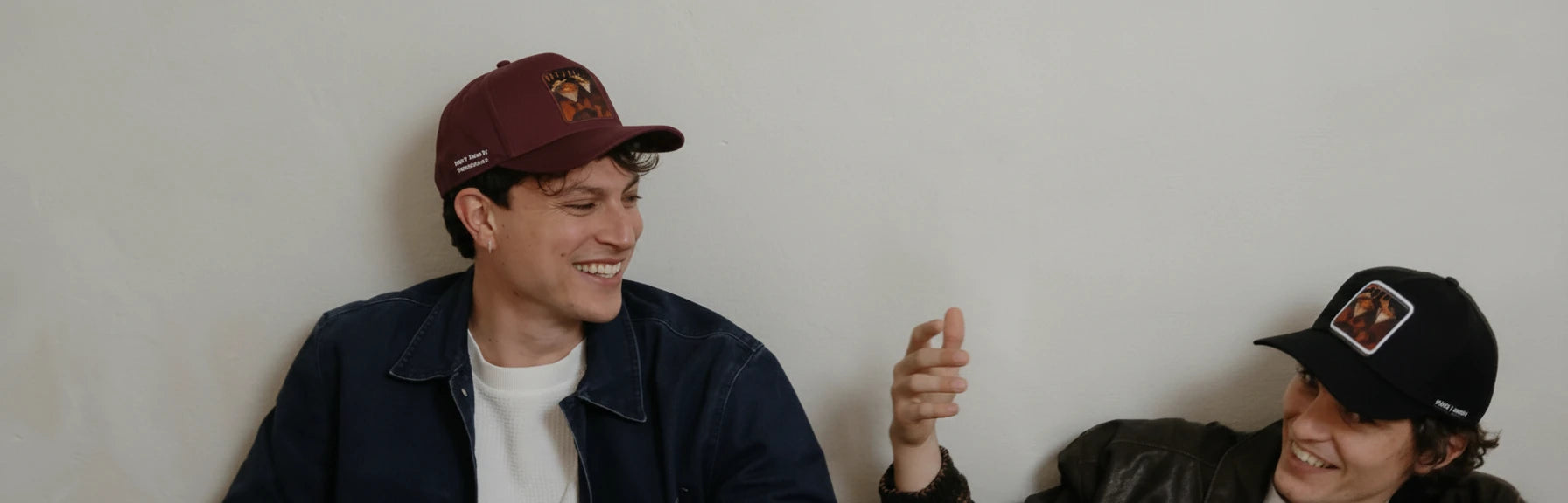 Two people sitting against a plain wall, wearing caps and jackets.