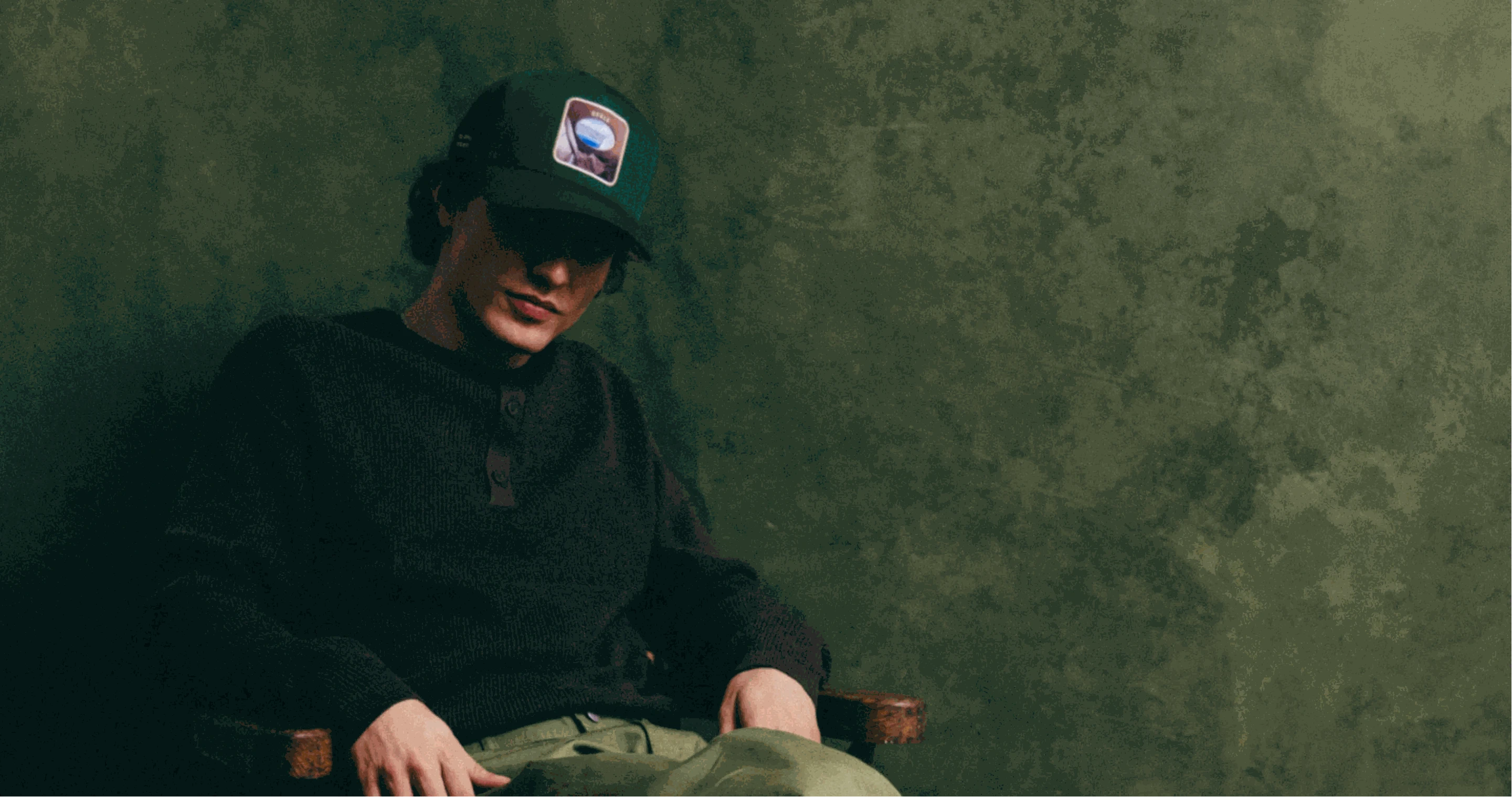 Man wearing a dark green Ovals Baseball Cap with embroidered patch and casual outfit sitting against a green textured background